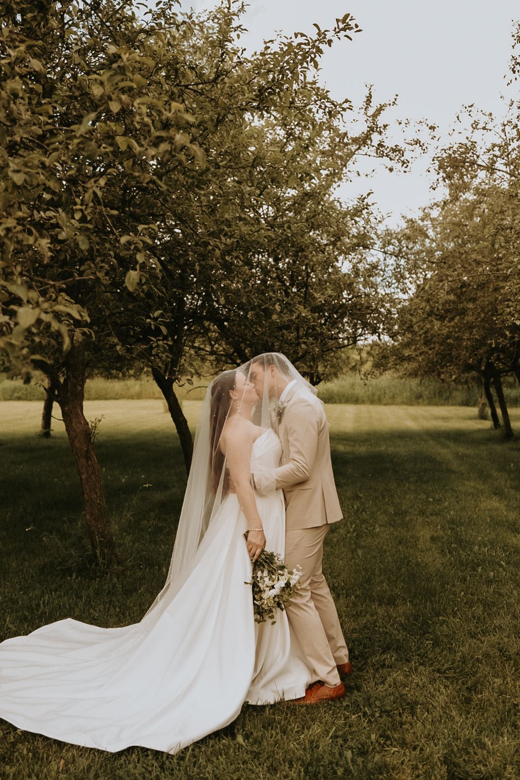 Couple shares intimate moment under trees with flowing white wedding dress and veil.