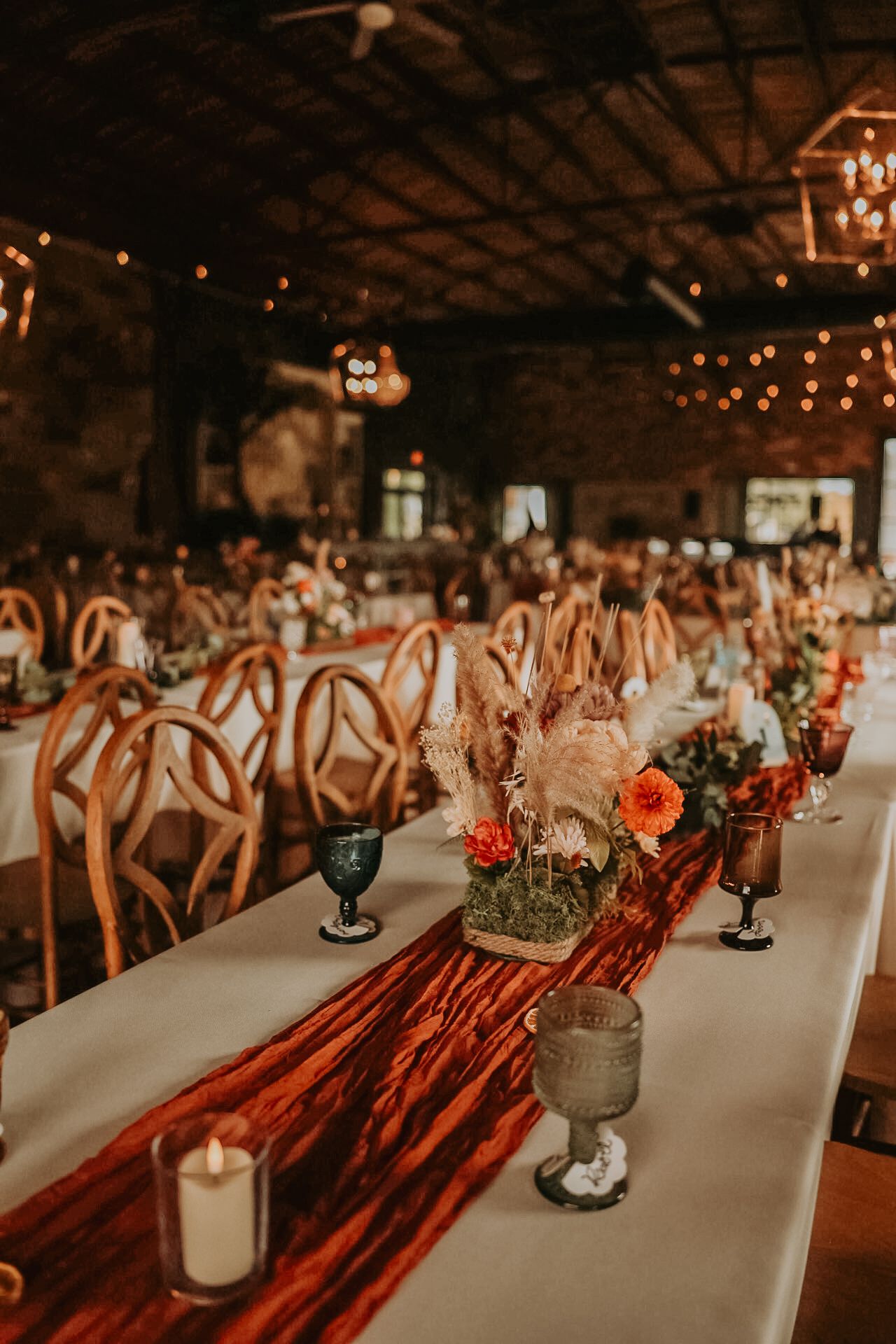 Rustic wedding reception venue with wooden chairs and red table runners decorated with floral centerpieces.