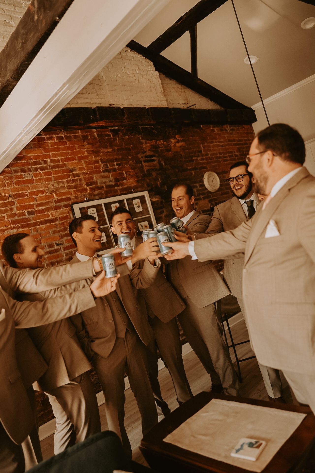 Wedding party in tan suits raising beer cans in a toast against a brick wall backdrop.