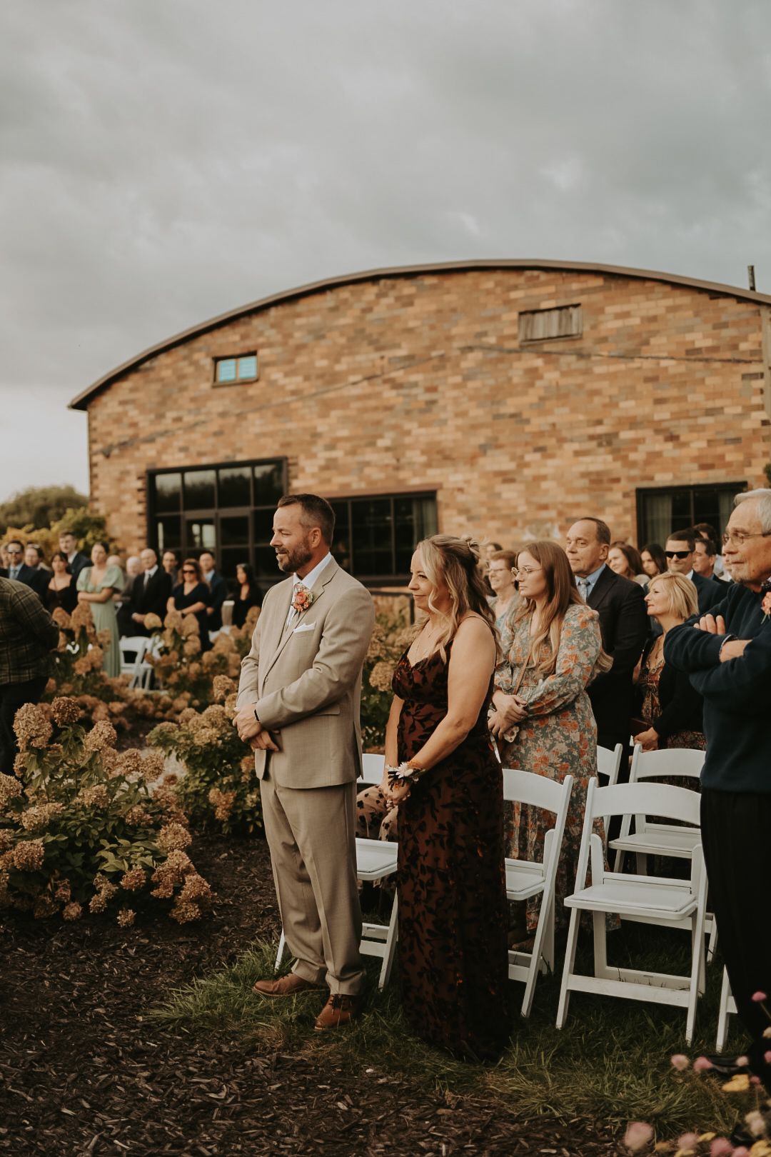 Wedding guests stand during ceremony in front of elegant brick building.