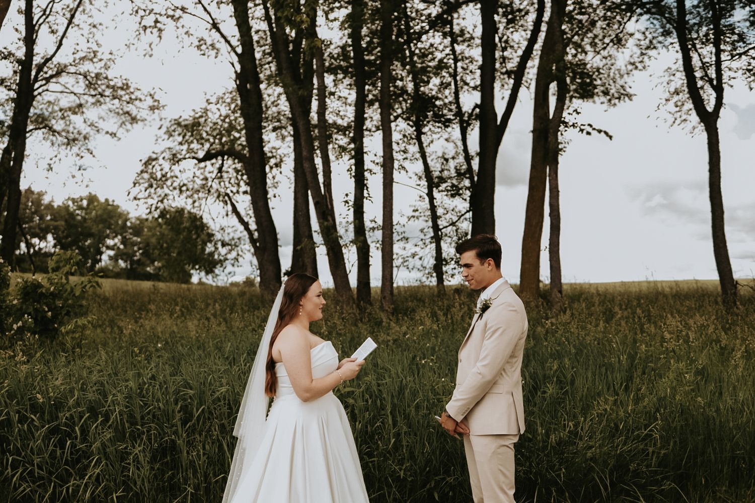 Couple reads vows to each other in a scenic field with tall trees in the background.