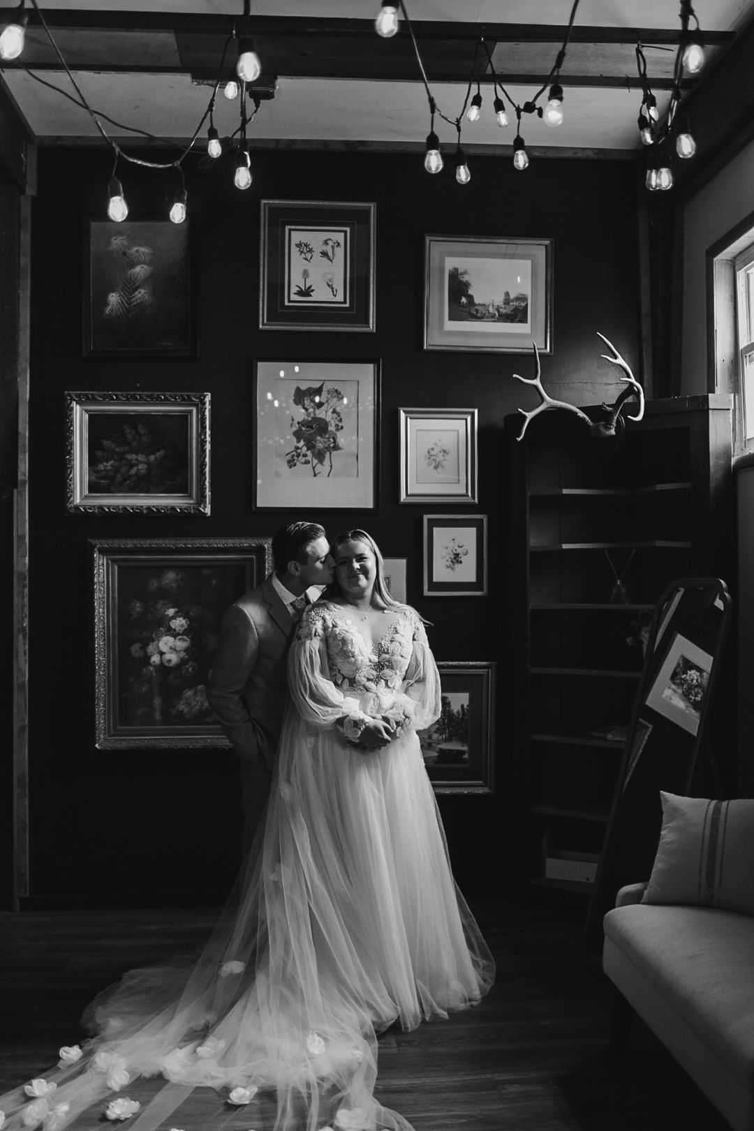 Moody black and white wedding photo with string lights and gallery wall backdrop.