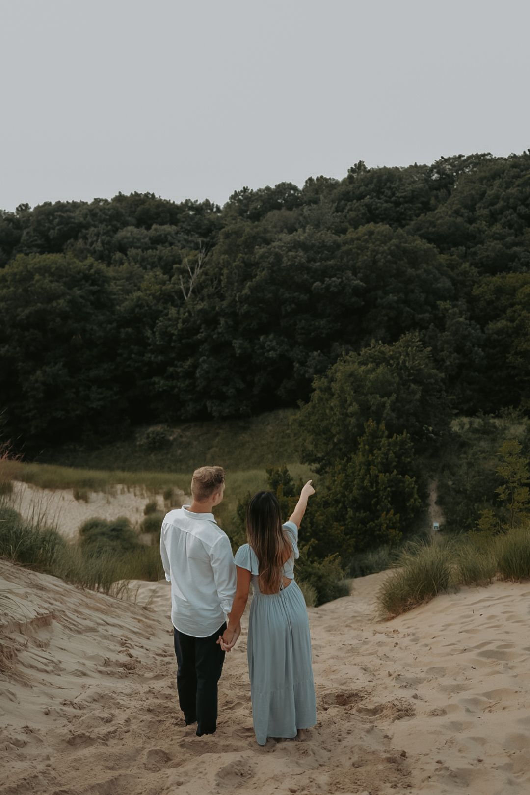 A couple stands together on sand dunes overlooking a forested area.