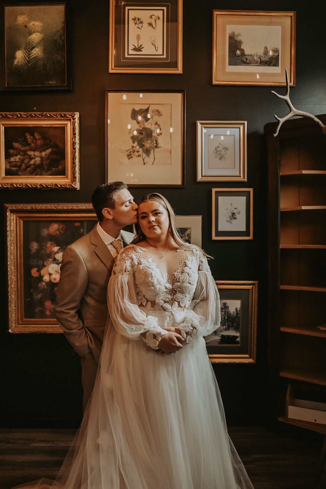 Couple shares romantic moment in dimly lit room with vintage artwork and frames on walls.