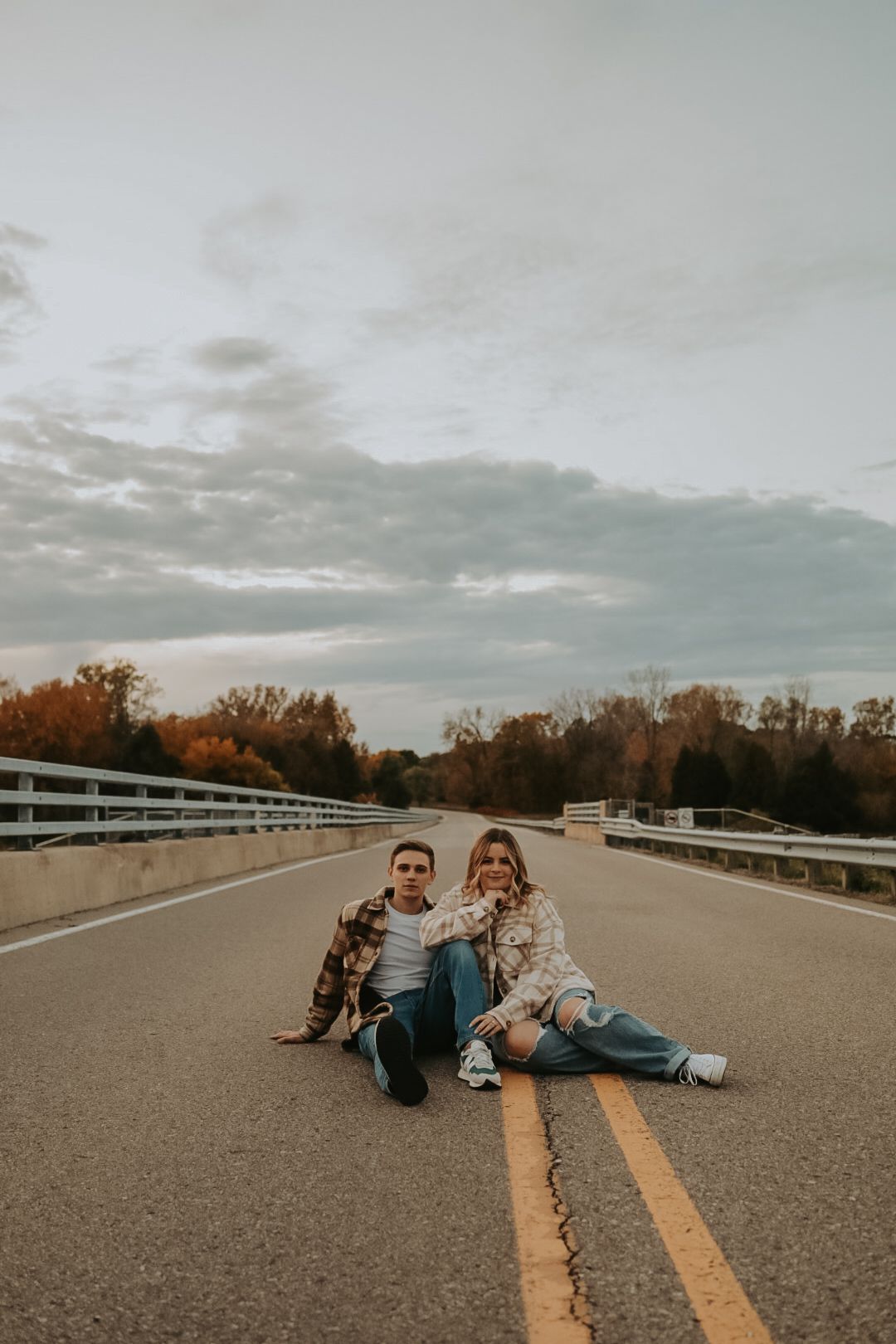 Two people in casual wear sitting on yellow lines of empty road during sunset.