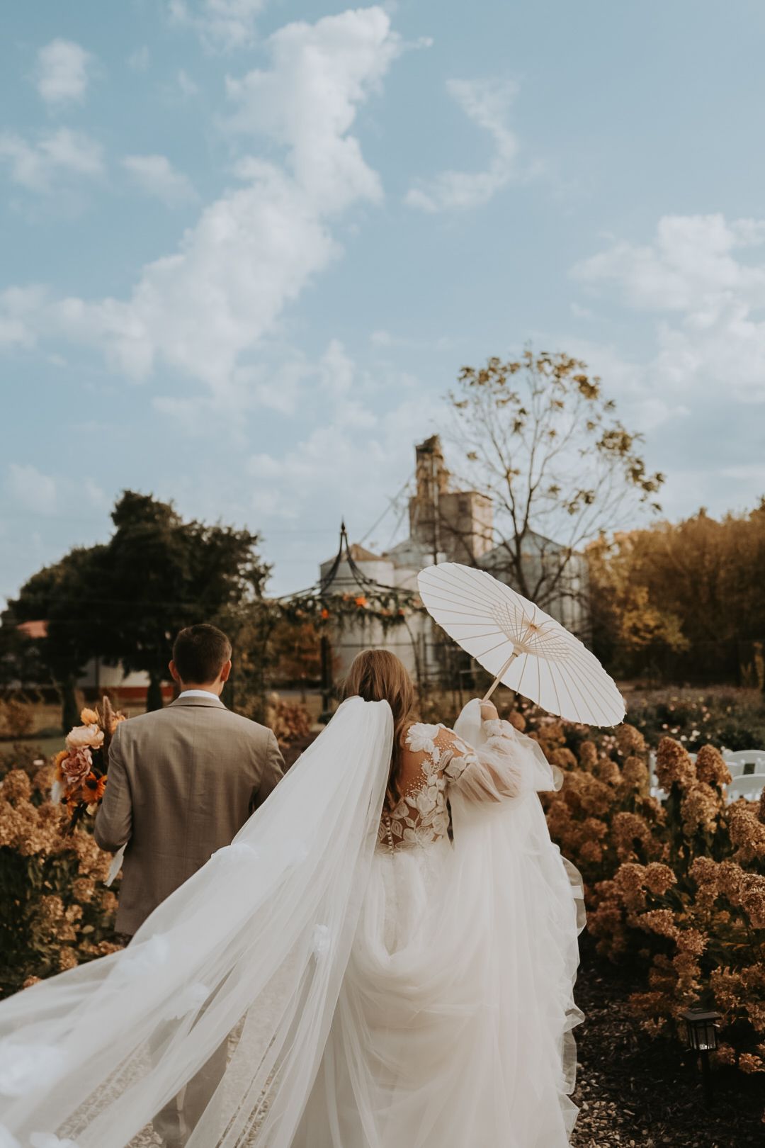 A couple walks through a garden holding a white parasol with a church spire visible in the background.
