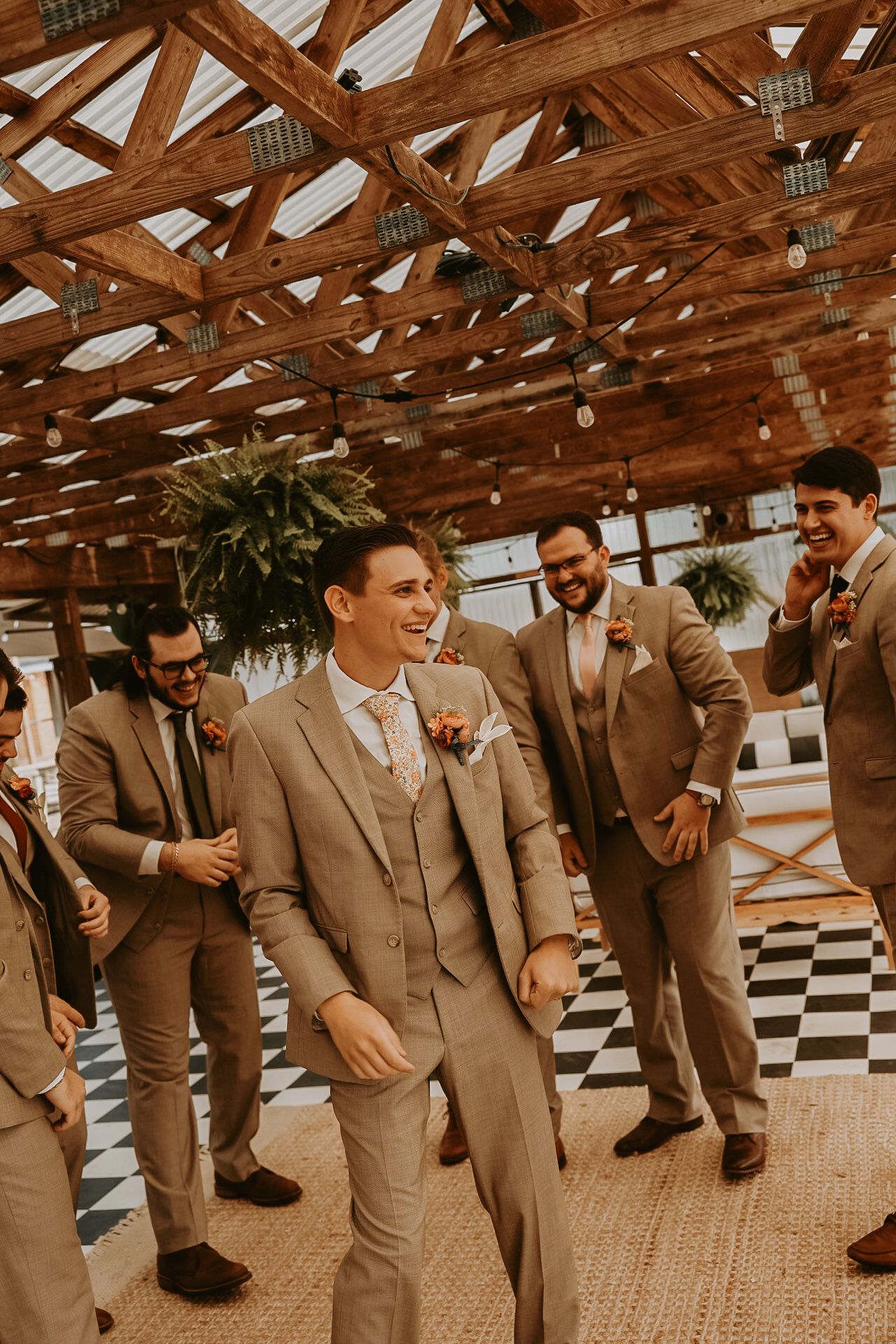 Groomsmen in tan suits pose playfully on a checkered floor under wooden beams.