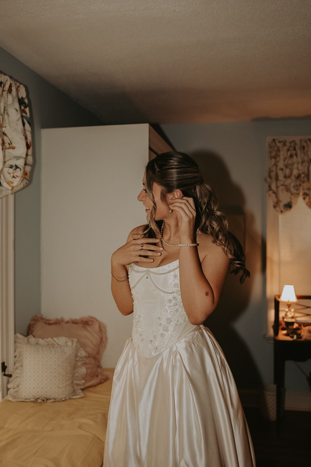 Two figures in white dresses share an intimate moment in a warmly lit bedroom setting.