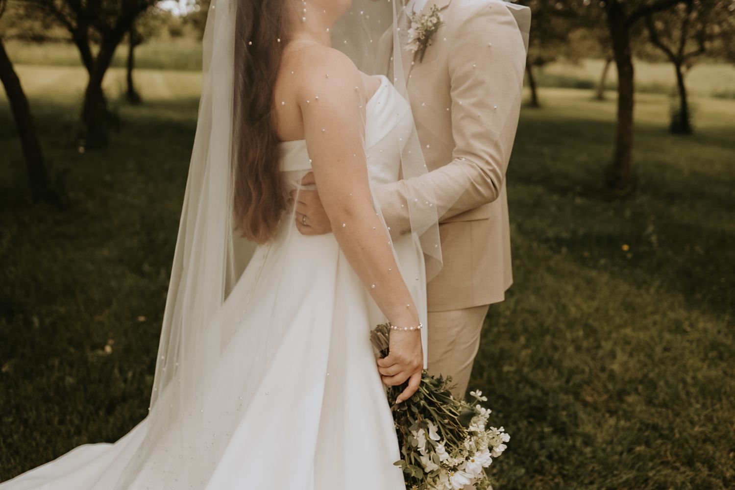 Couple in elegant wedding attire poses together holding a bouquet in an orchard setting.