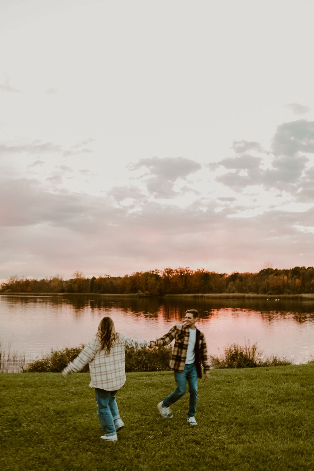 Two people playfully walk on grass near a lake during a golden sunset.