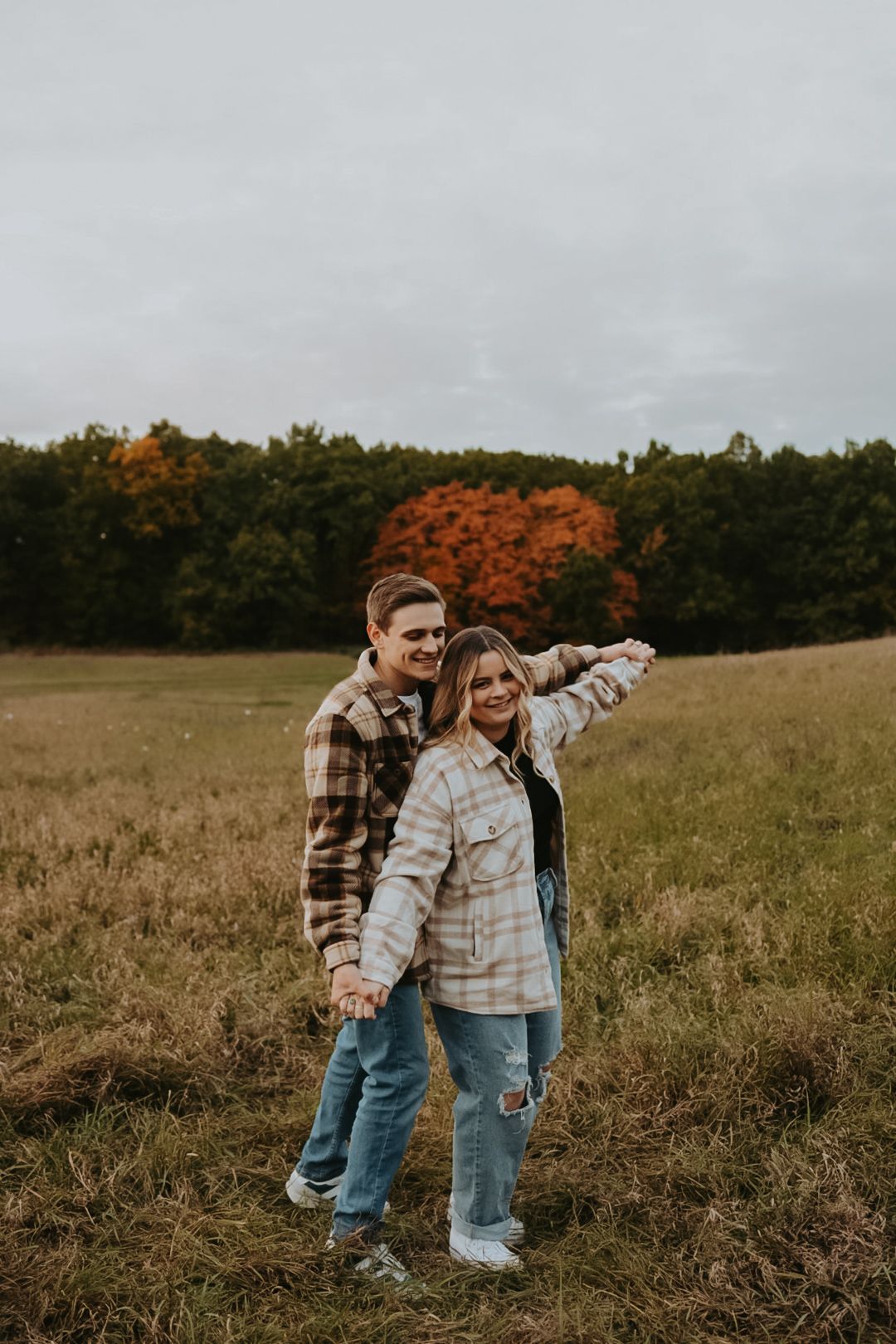 A couple dances and laughs together in a rustic field with fall foliage in background.