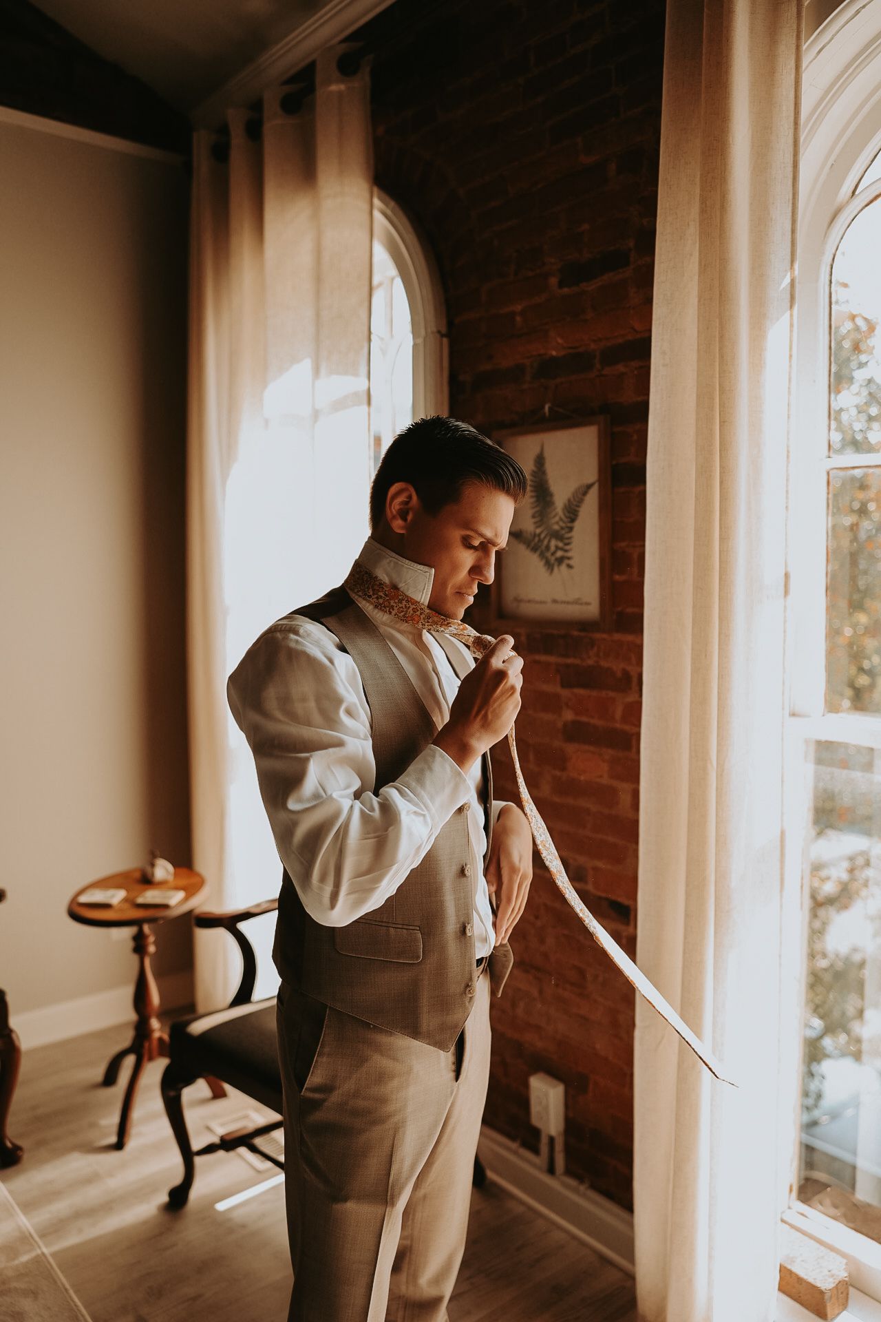 A person in formal attire adjusts their tie while looking out a window with exposed brick walls.