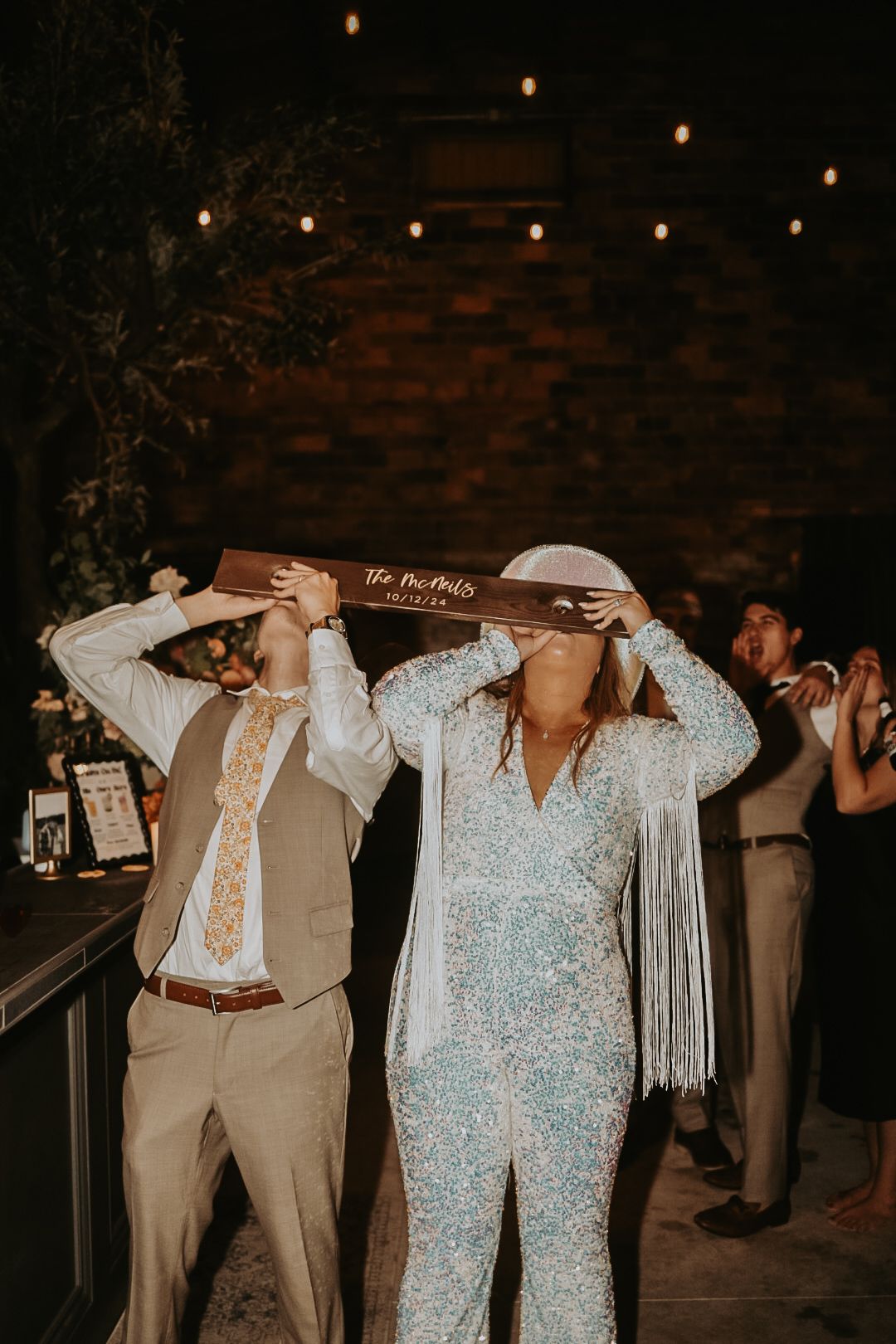 Wedding guests taking shots at a dimly lit reception with string lights and brick wall backdrop.