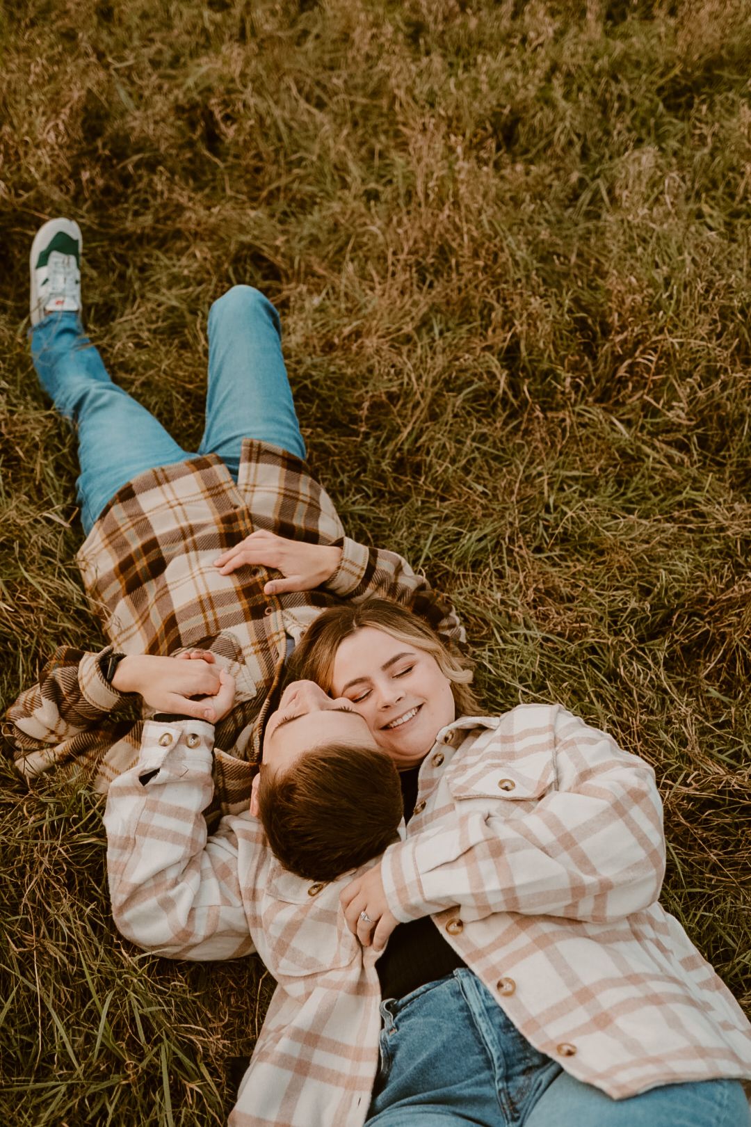 Two people in matching plaid shirts lay together in tall grass during autumn.