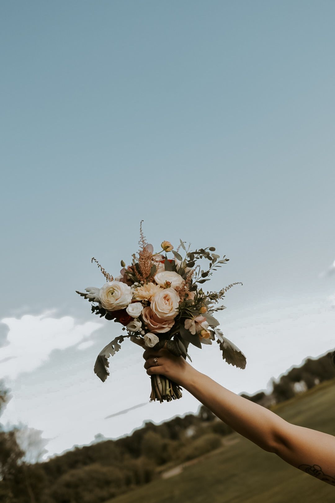 Hand holding elegant bouquet with roses and greenery against blue sky.