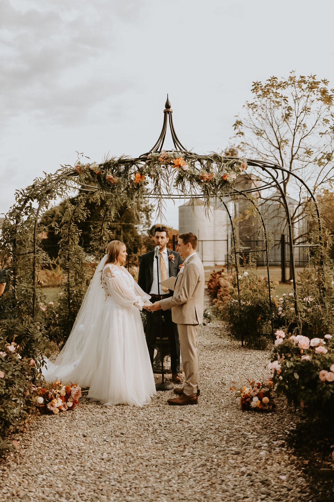 Wedding ceremony under metal gazebo decorated with autumn flowers and greenery.