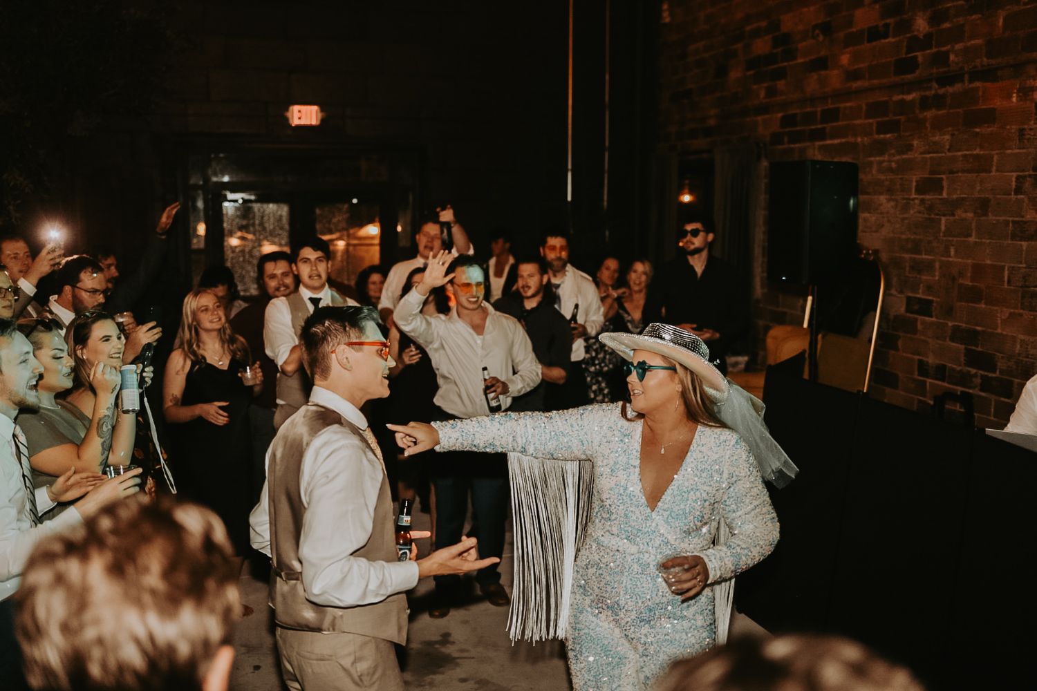 Lively wedding reception with guests dancing in a brick-walled venue with ambient lighting.