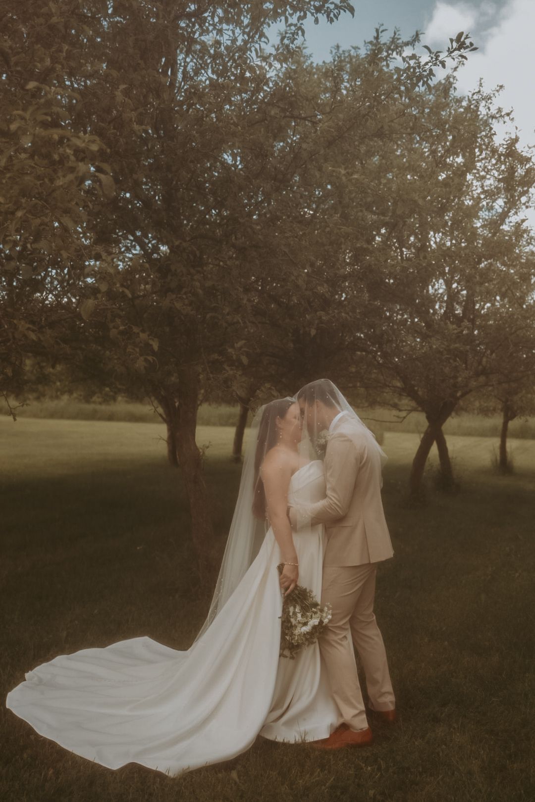 Romantic wedding portrait in a dreamy orchard setting with flowing white dress and veil.
