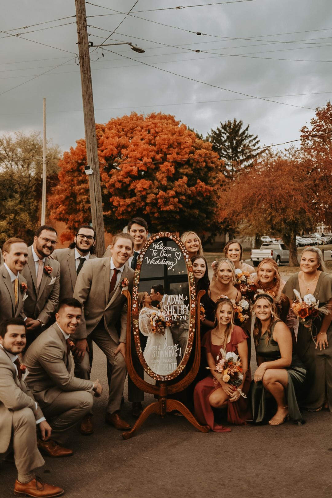 Large wedding party poses with decorative signs against backdrop of vibrant fall foliage.