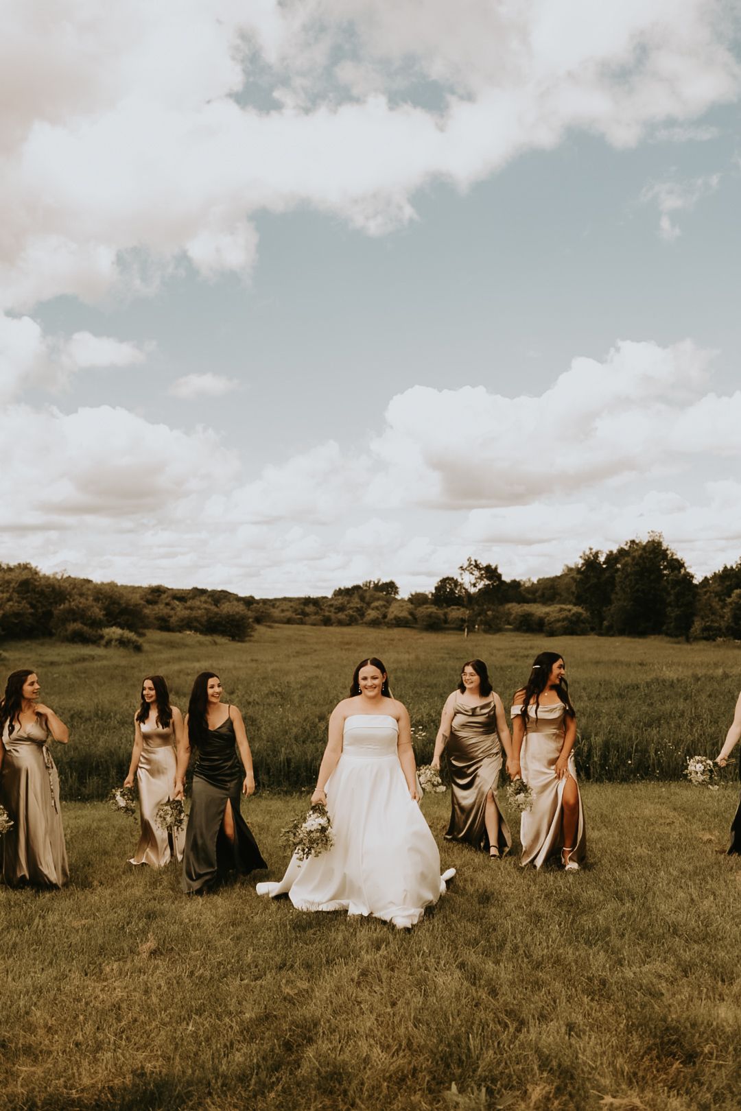 Bridal party sits together on grassy hillside wearing neutral colored dresses.