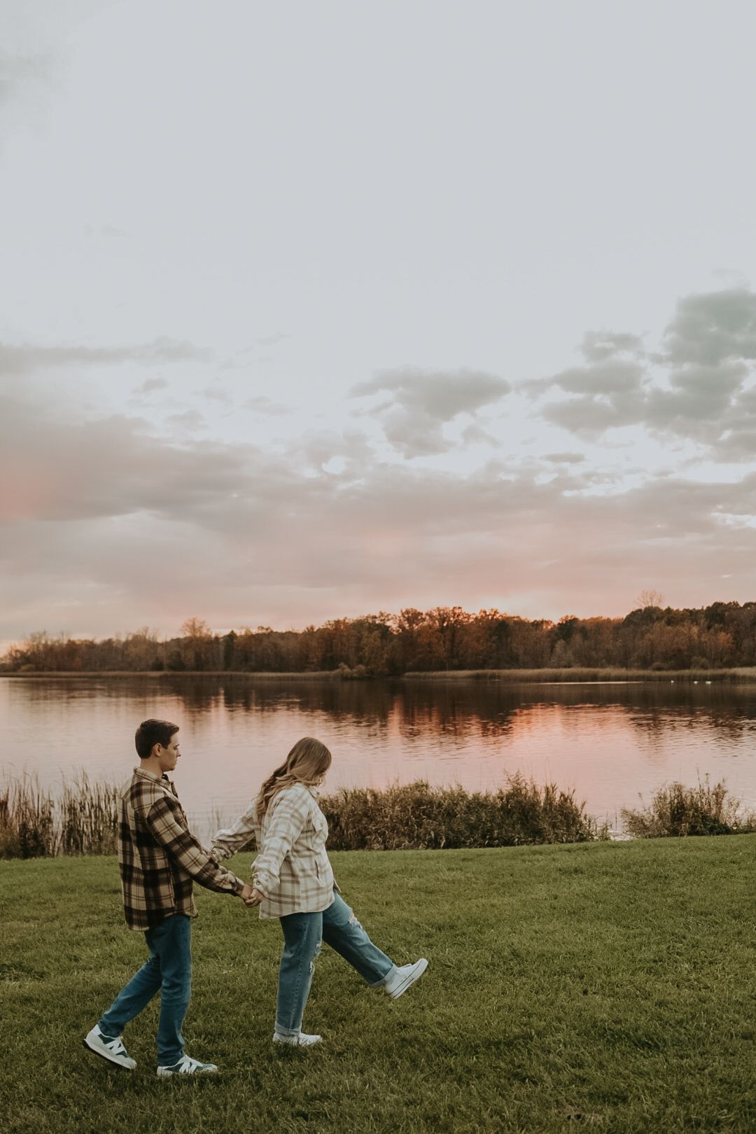 A couple strolls along a lakeside path during sunset with autumn trees reflecting on the calm water.