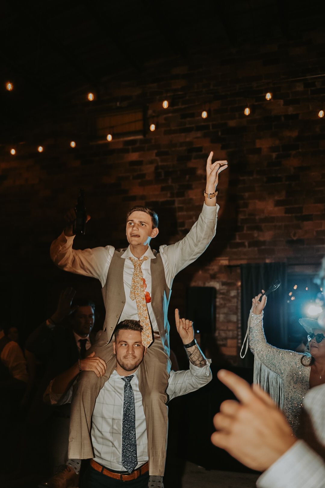 Wedding guests dancing enthusiastically under string lights at an indoor reception.