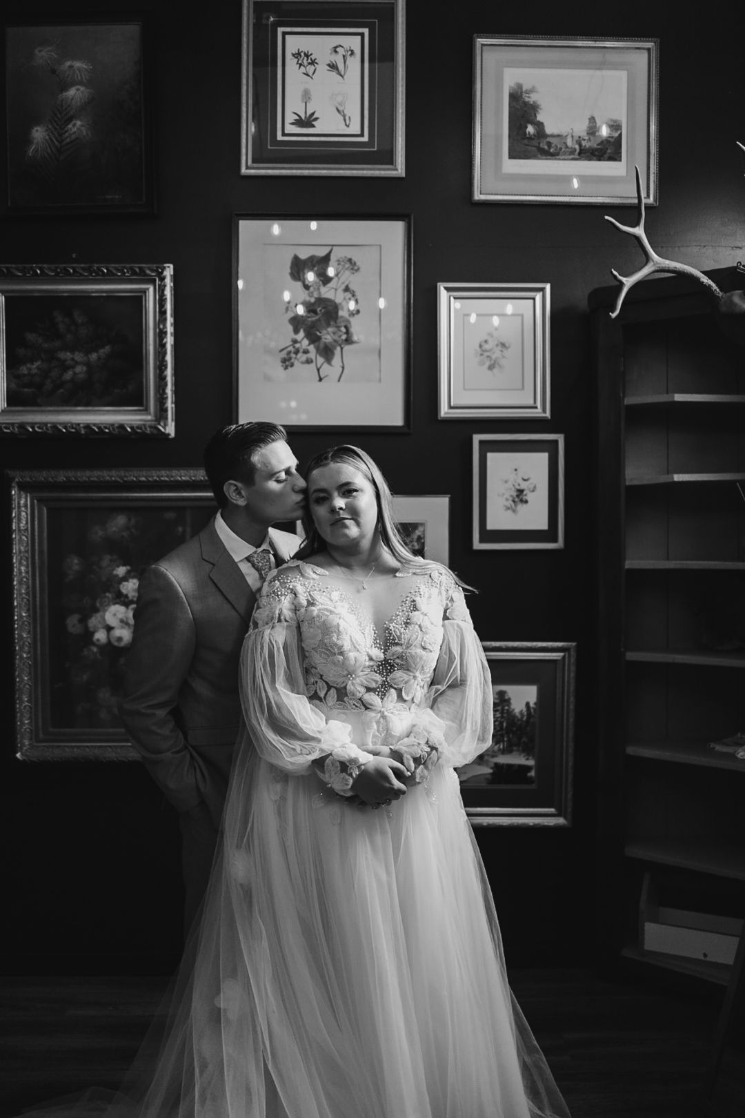 Black and white portrait of couple embracing in elegant room with framed artwork.