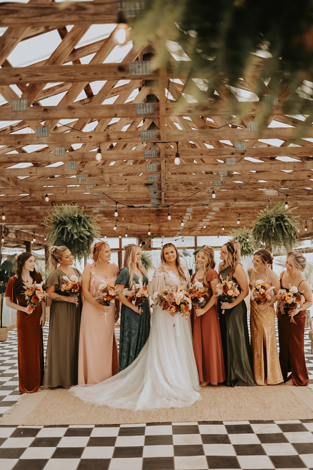 Wedding party in earth-toned dresses standing under wooden beams on checkered floor.