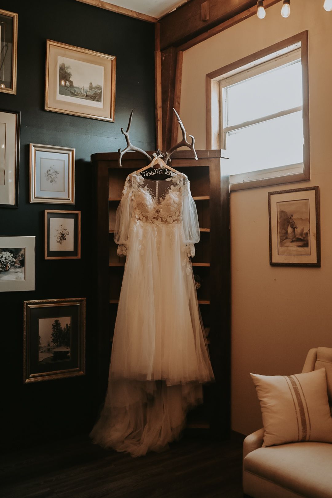 A delicate white wedding dress with lace details hangs in a corner room decorated with vintage framed artwork.