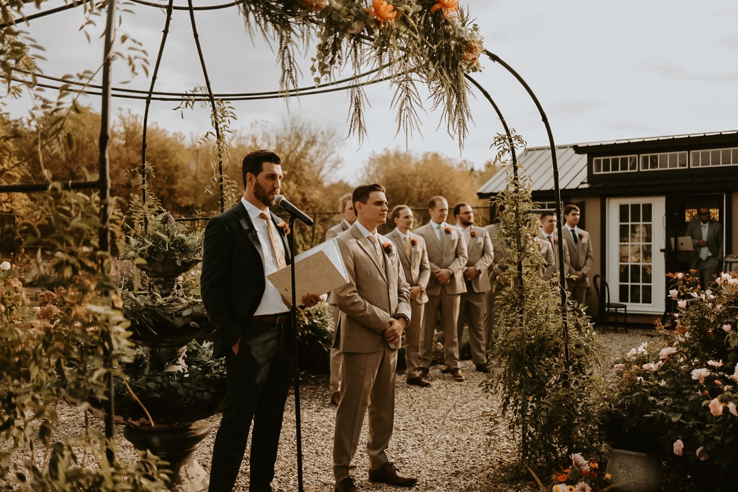 Outdoor autumn wedding ceremony with floral arch and guests gathered in garden setting.
