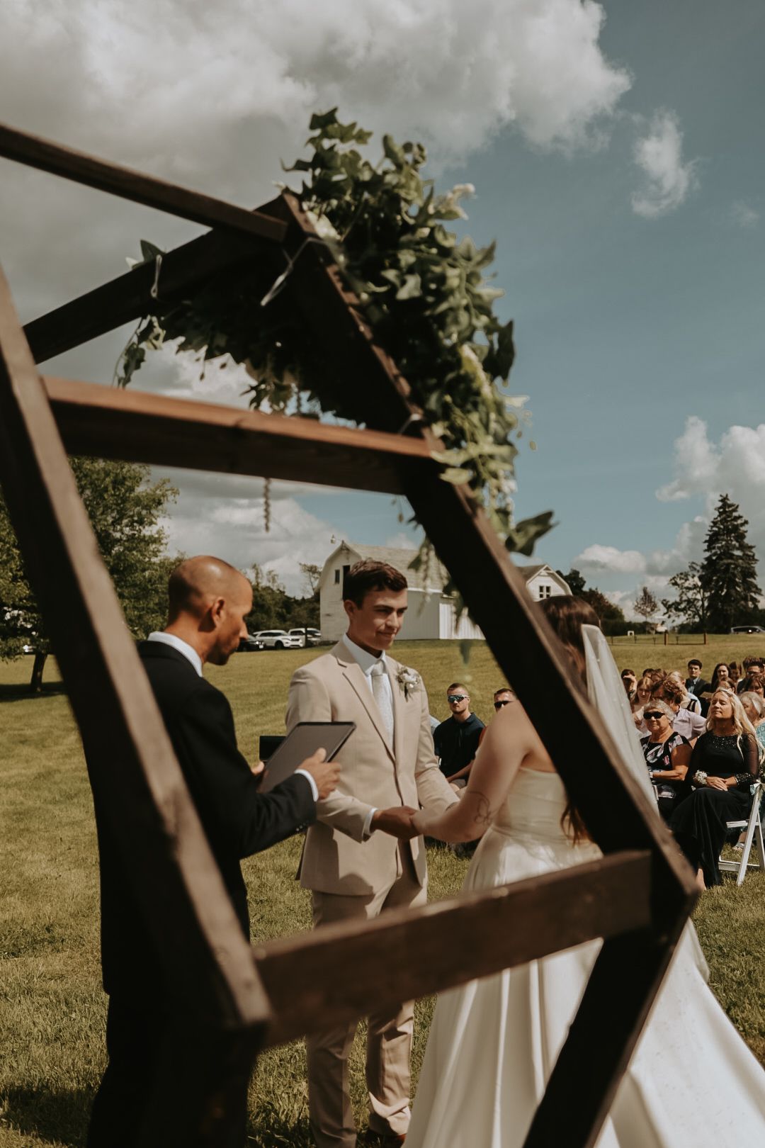 Outdoor wedding ceremony viewed through wooden arch frame with guests seated.