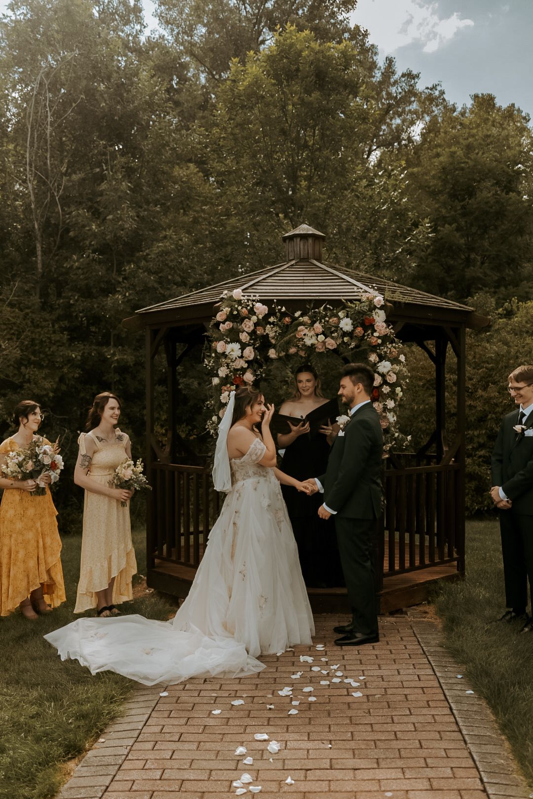 An outdoor wedding ceremony takes place under a floral arch on a stone patio.