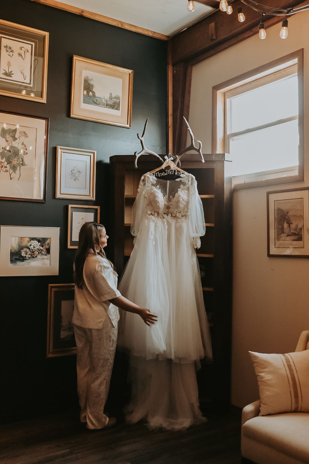 An ethereal wedding dress hangs from antlers in a cozy room with vintage artwork and a beige sofa.