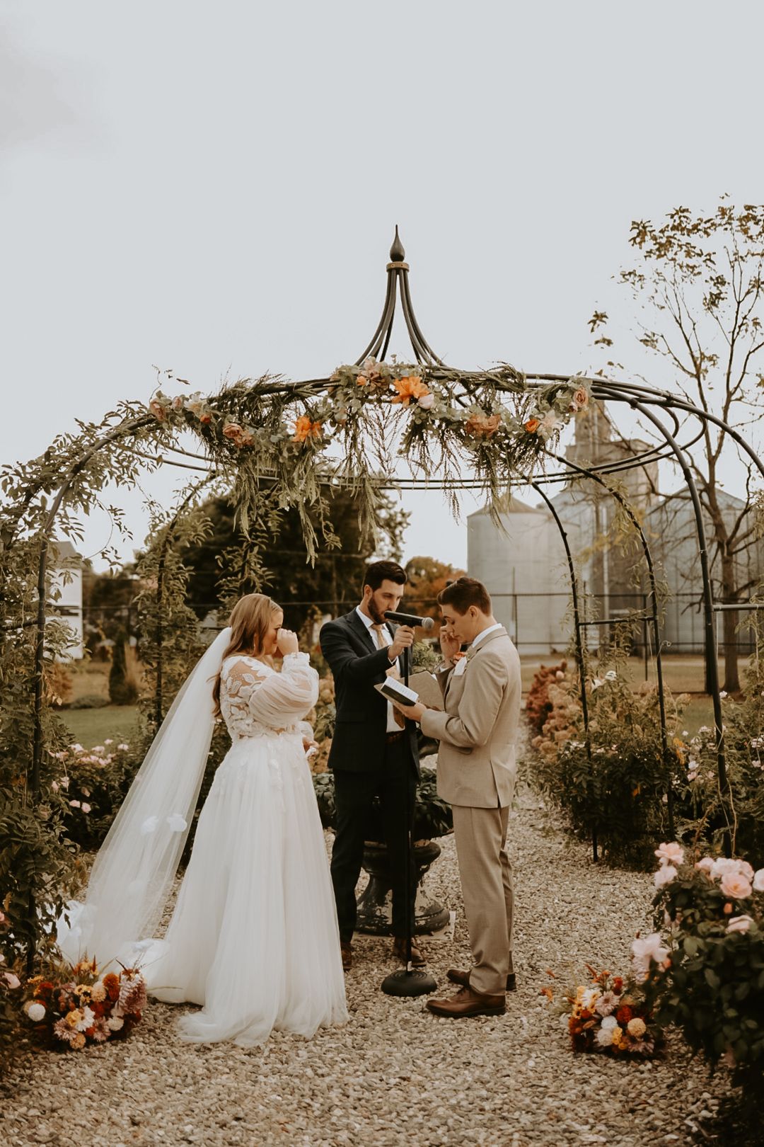 Autumn wedding ceremony under metal gazebo with floral decorations and gathered guests.