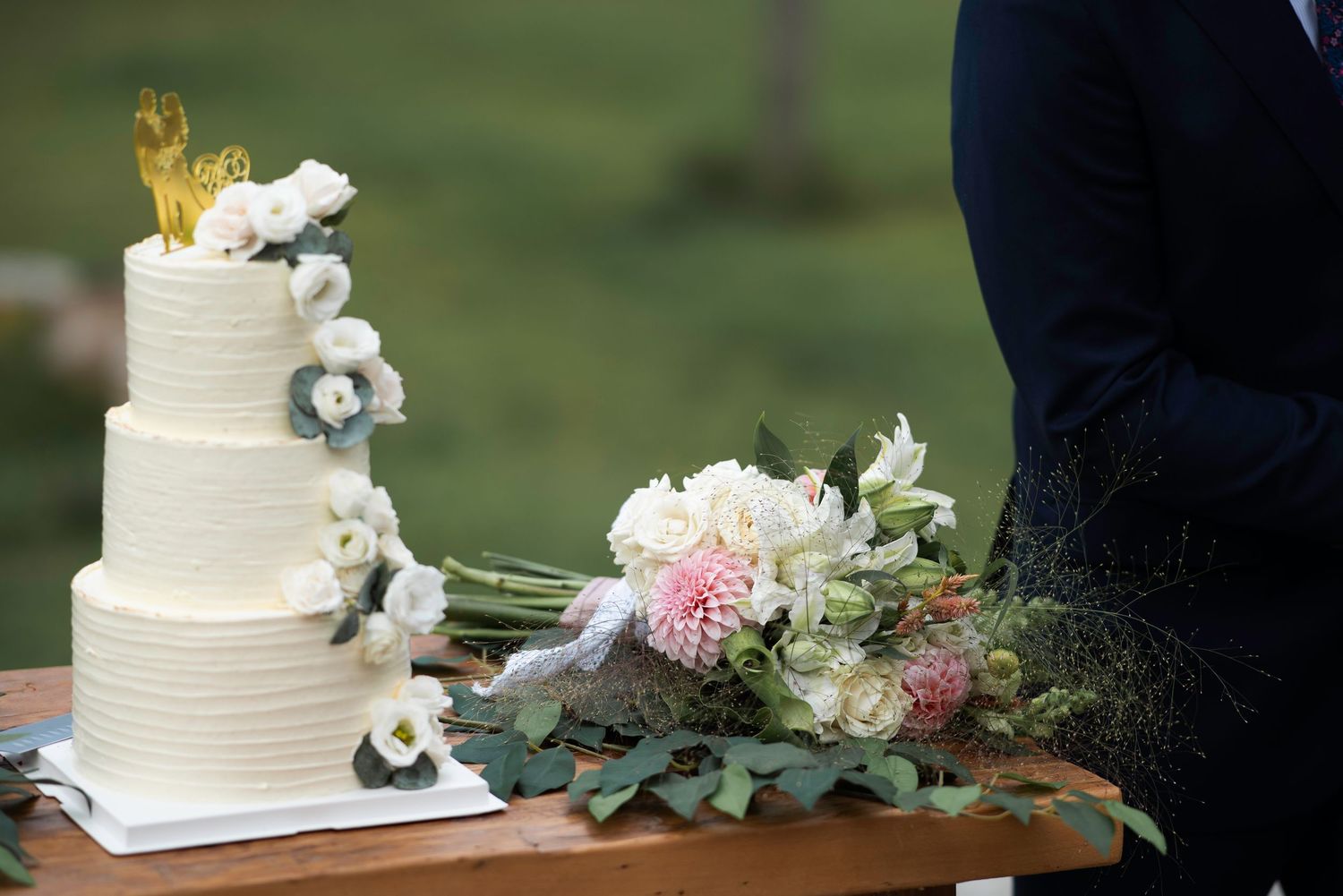 White wedding cakes decorated with fresh flowers and greenery displayed on wooden tables at an outdoor reception.