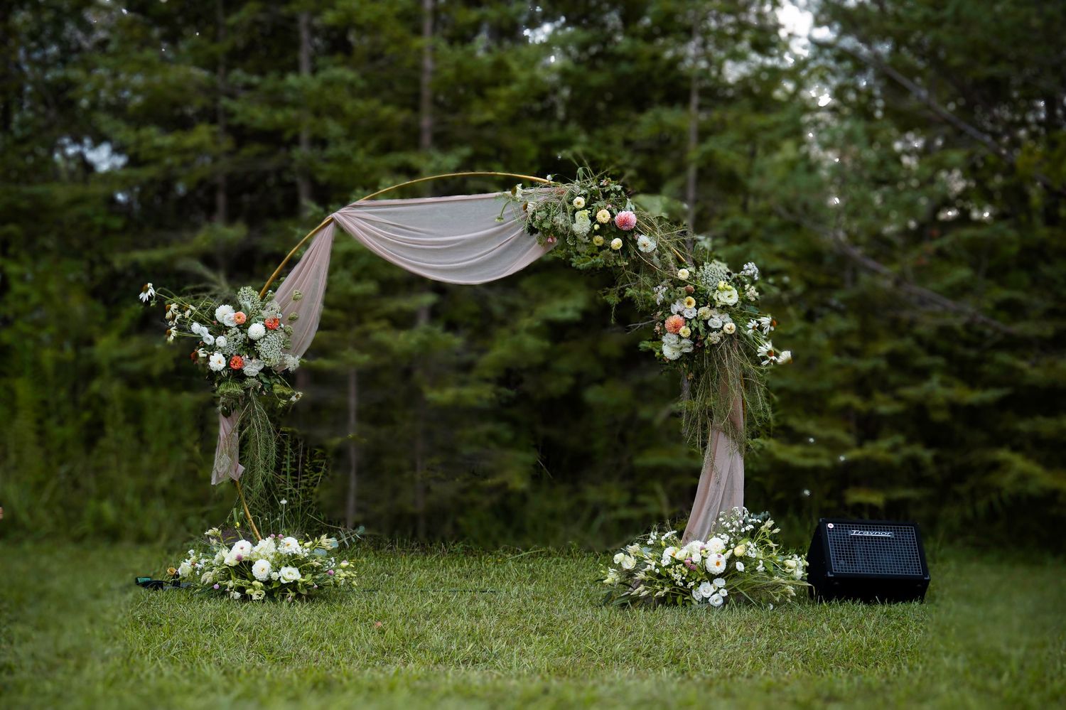 A rustic outdoor wedding ceremony setup with a white draped arch and floral arrangements in front of evergreen trees.