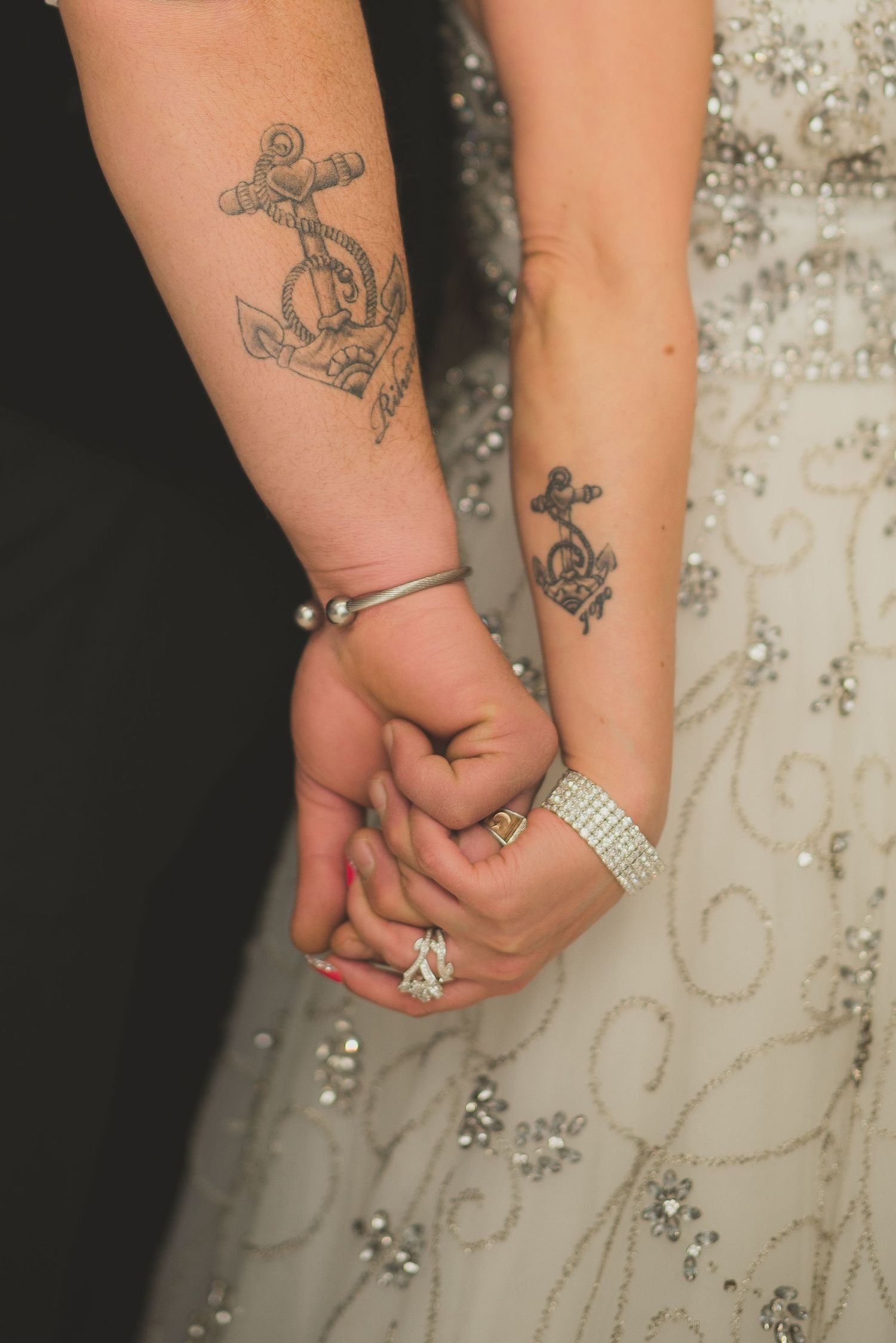 Close-up of clasped hands showing wedding rings and tattoos against an embellished white dress.