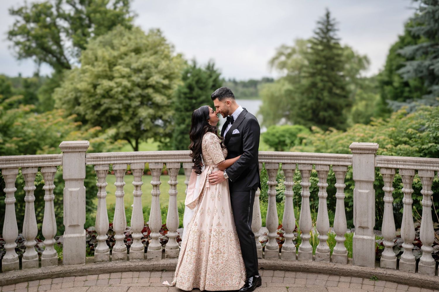 A romantic wedding couple embraces on an elegant stone balcony surrounded by lush greenery.