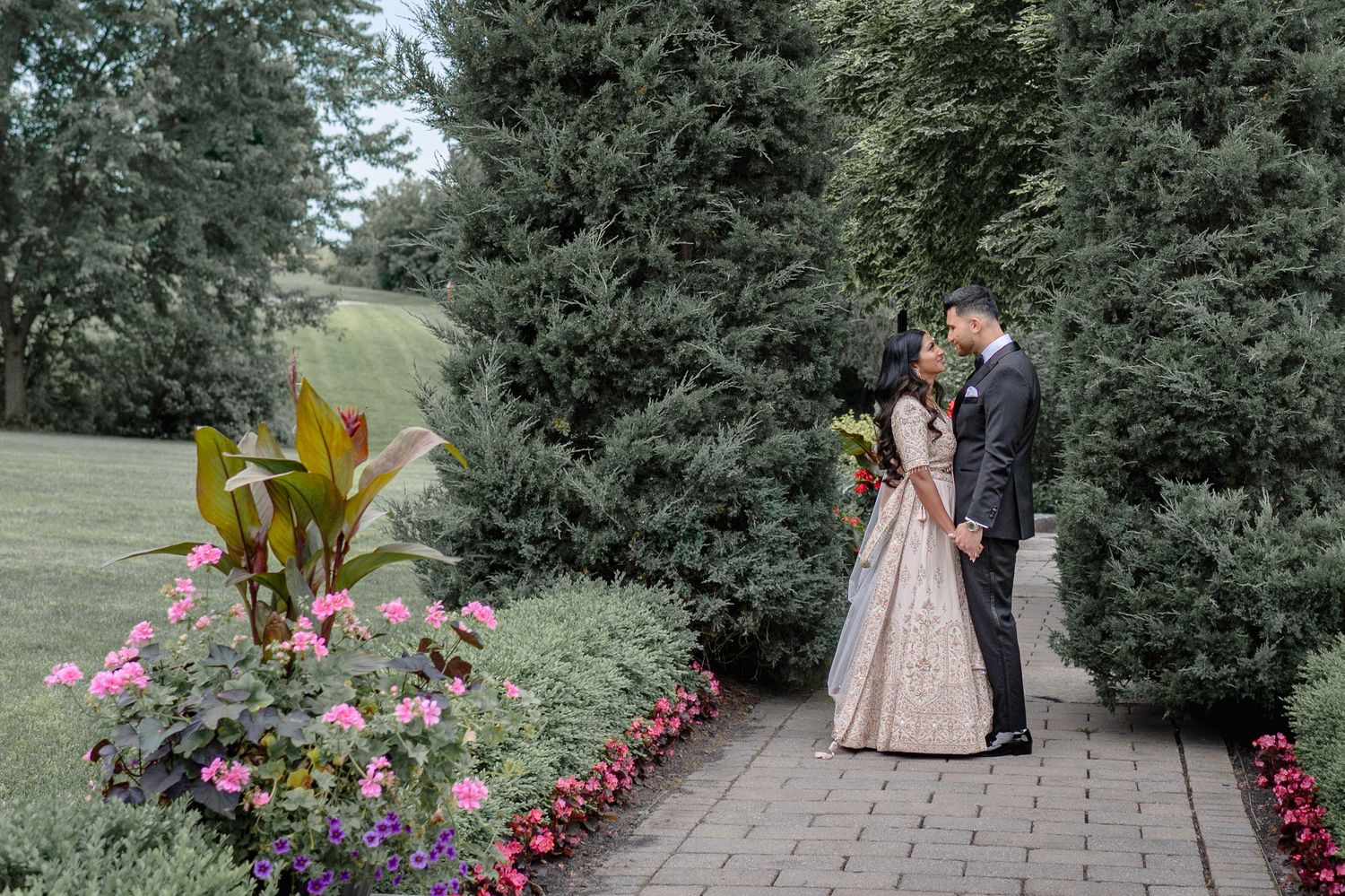 A couple shares a tender moment in a garden with colorful flowers and evergreen trees.