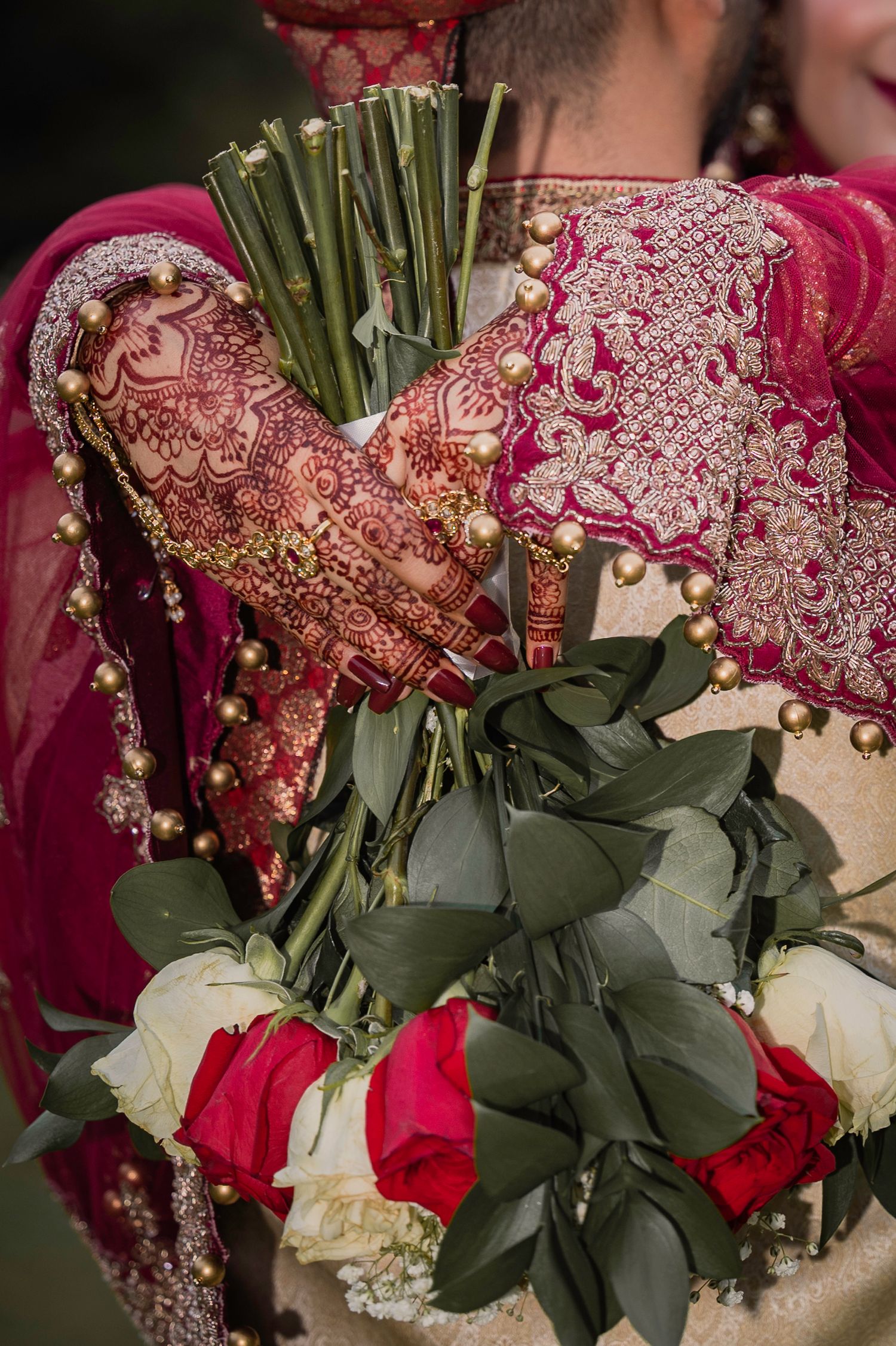 Elegant wedding bouquet with red roses and green foliage held against traditional Indian bridal attire with gold embroidery.