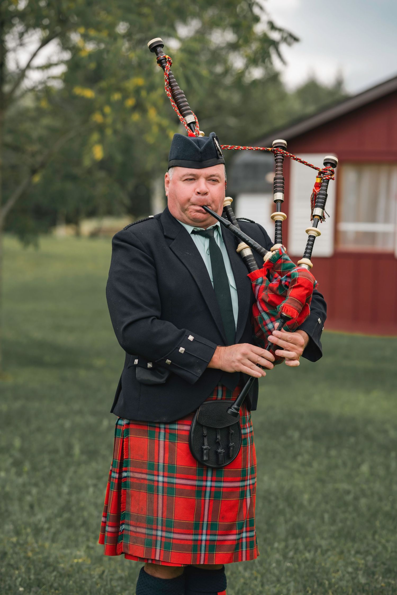 A bagpiper in traditional Scottish attire with red tartan kilt plays highland bagpipes outdoors near red buildings.