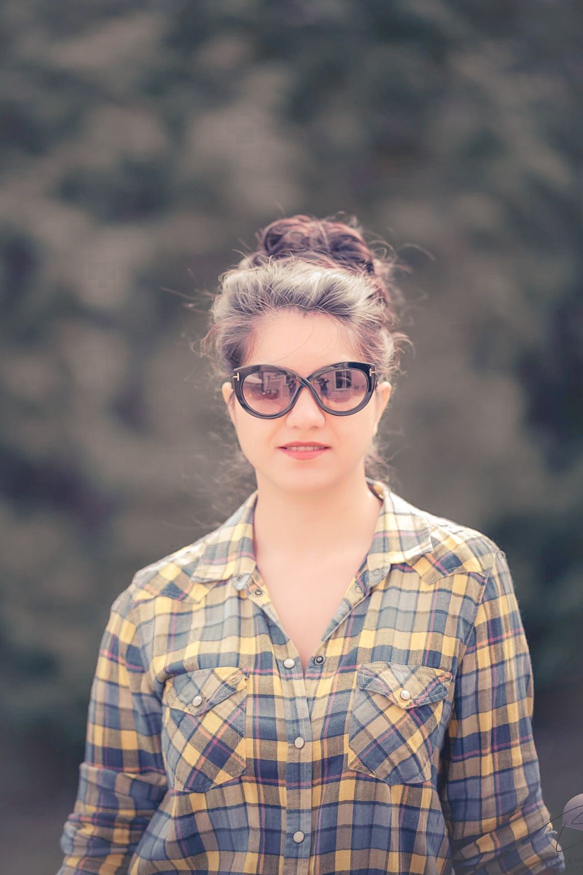 A person in a yellow plaid flannel shirt and cat-eye glasses smiles at the camera outdoors.