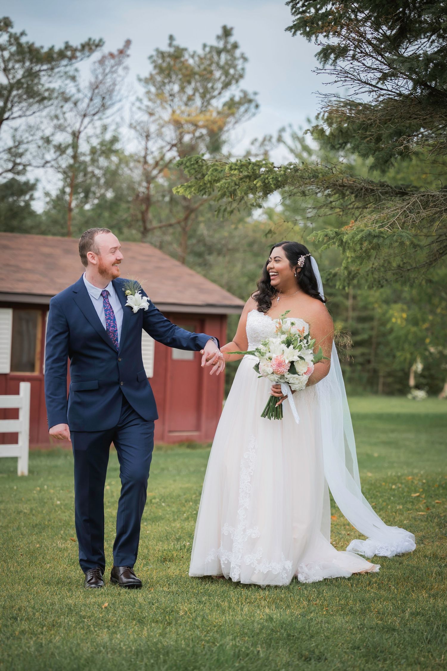 A couple in wedding attire holds hands while walking on grass with a rustic red barn in the background.