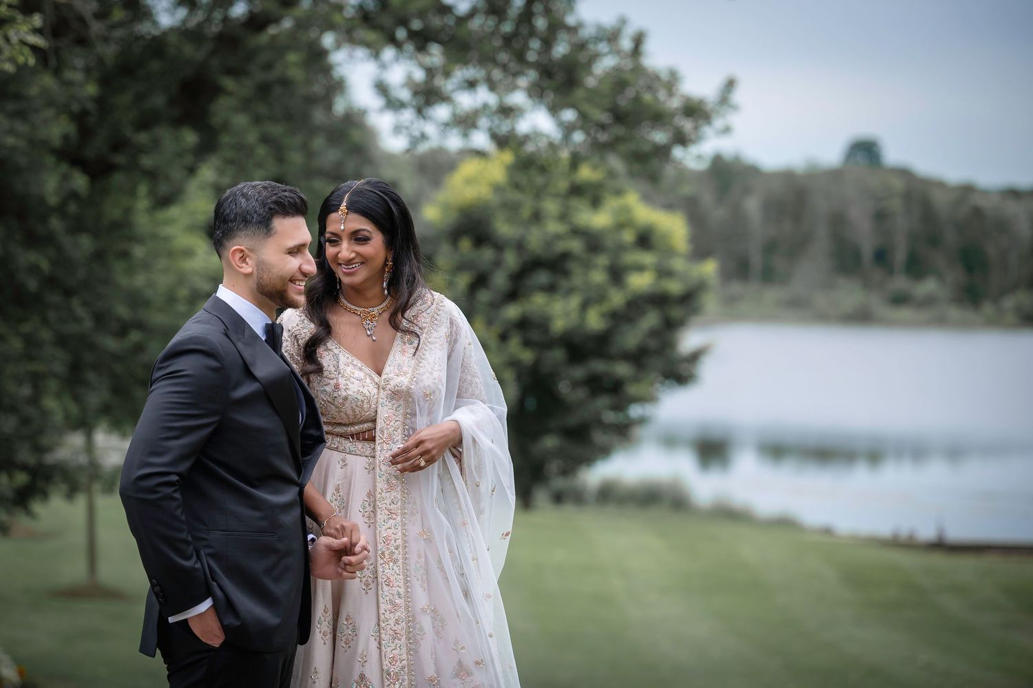 A couple in wedding attire poses by a serene lake with trees in the background.