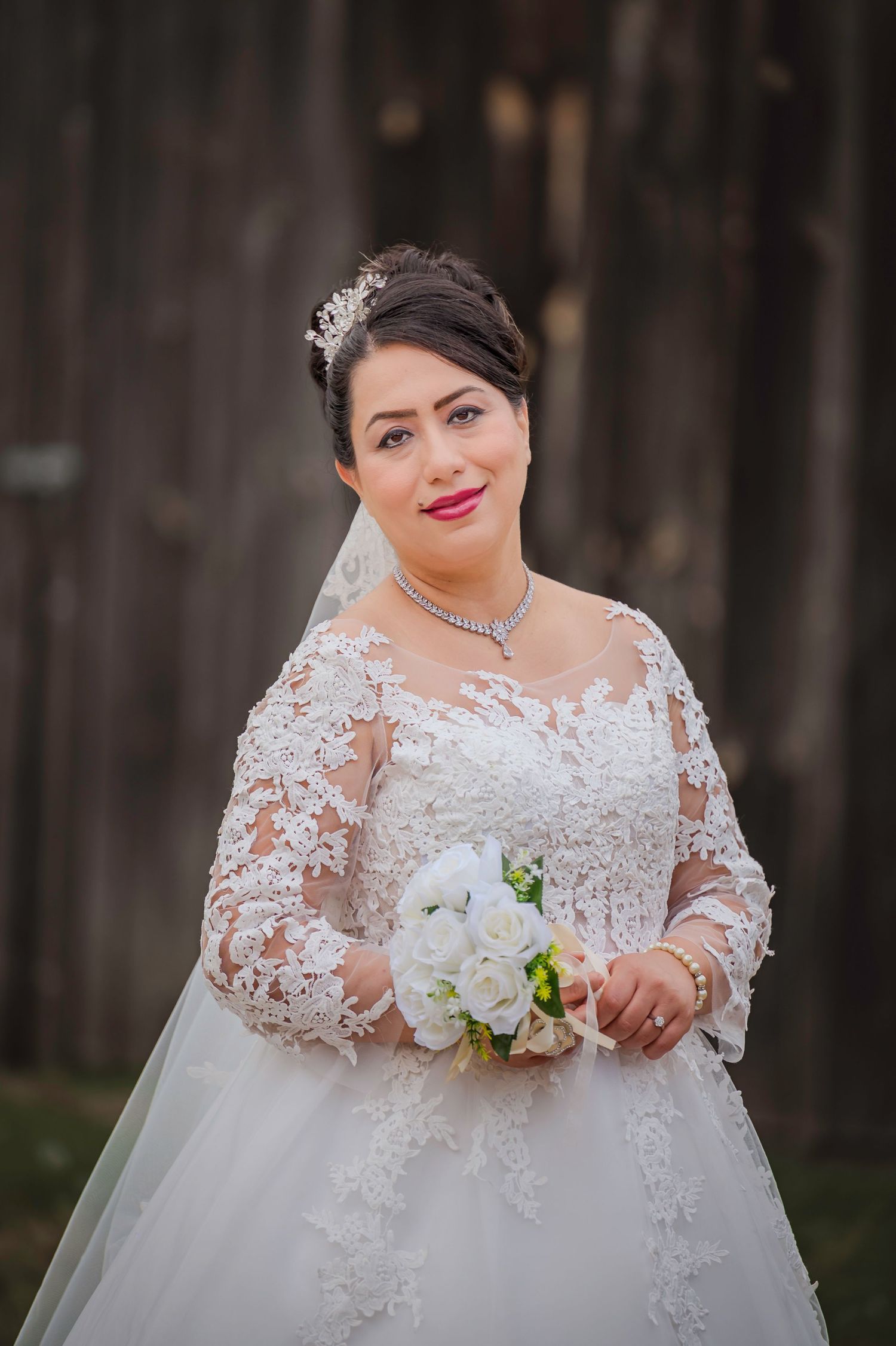 A bride in an elegant lace wedding gown holds a white bouquet against a dark background.