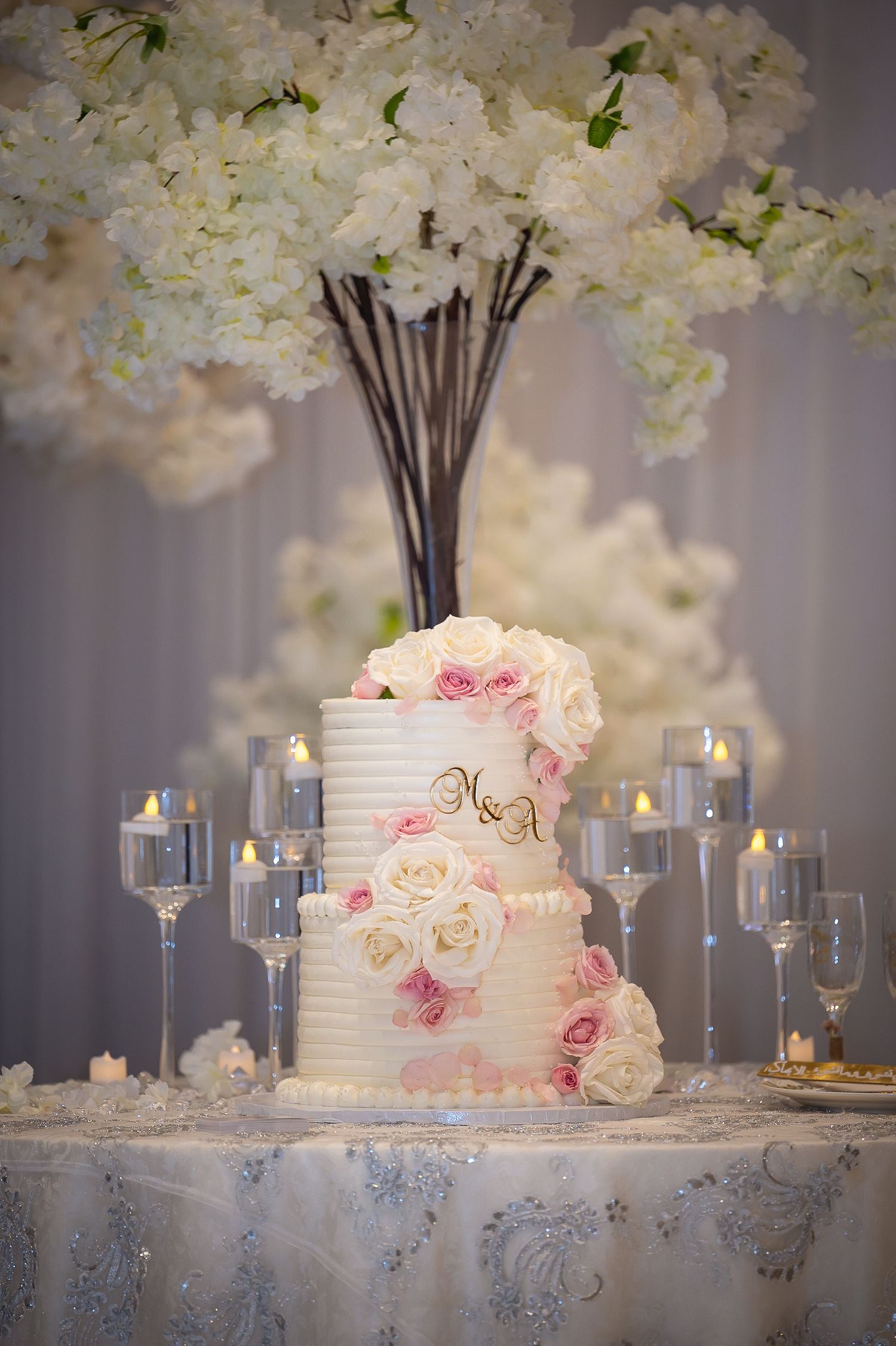 White wedding cake with pink roses displayed among white floral arrangements and candlelight.