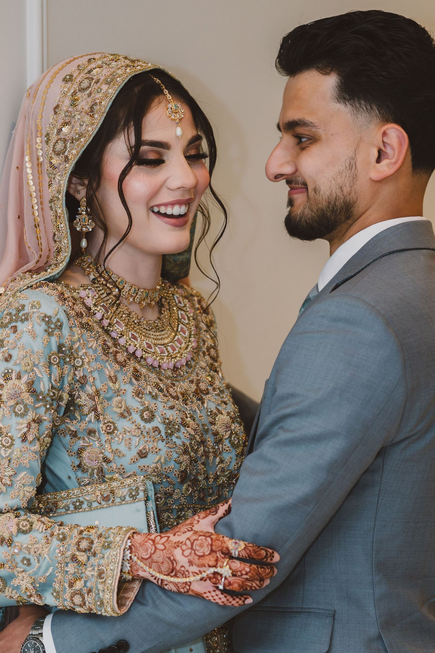 Bride in embellished blue lehenga and groom in grey suit share tender moments during their wedding photoshoot.