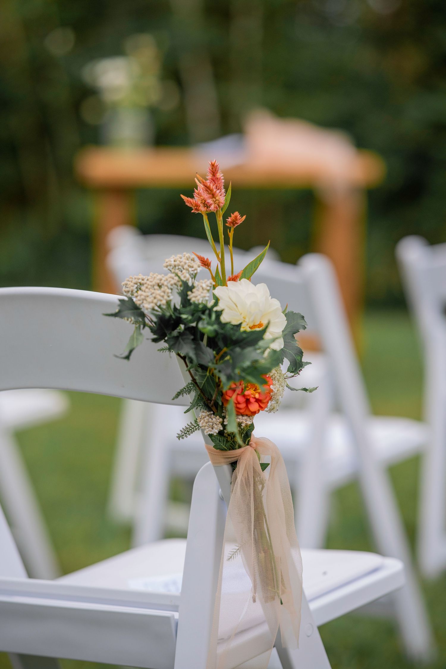 White folding chairs adorned with delicate floral arrangements line an outdoor wedding ceremony aisle.