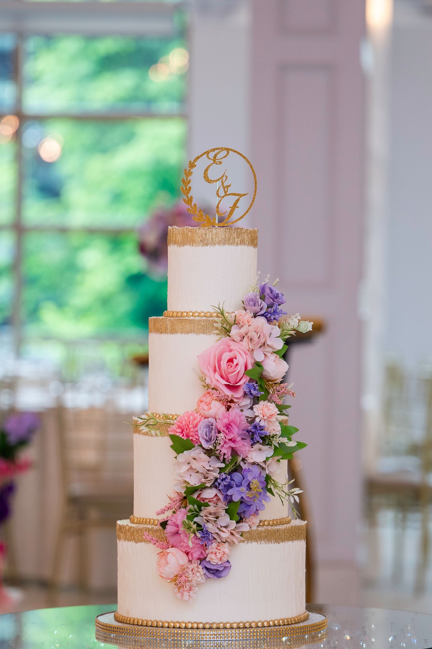 Three-tiered white wedding cakes decorated with pink and purple flowers and gold accents.