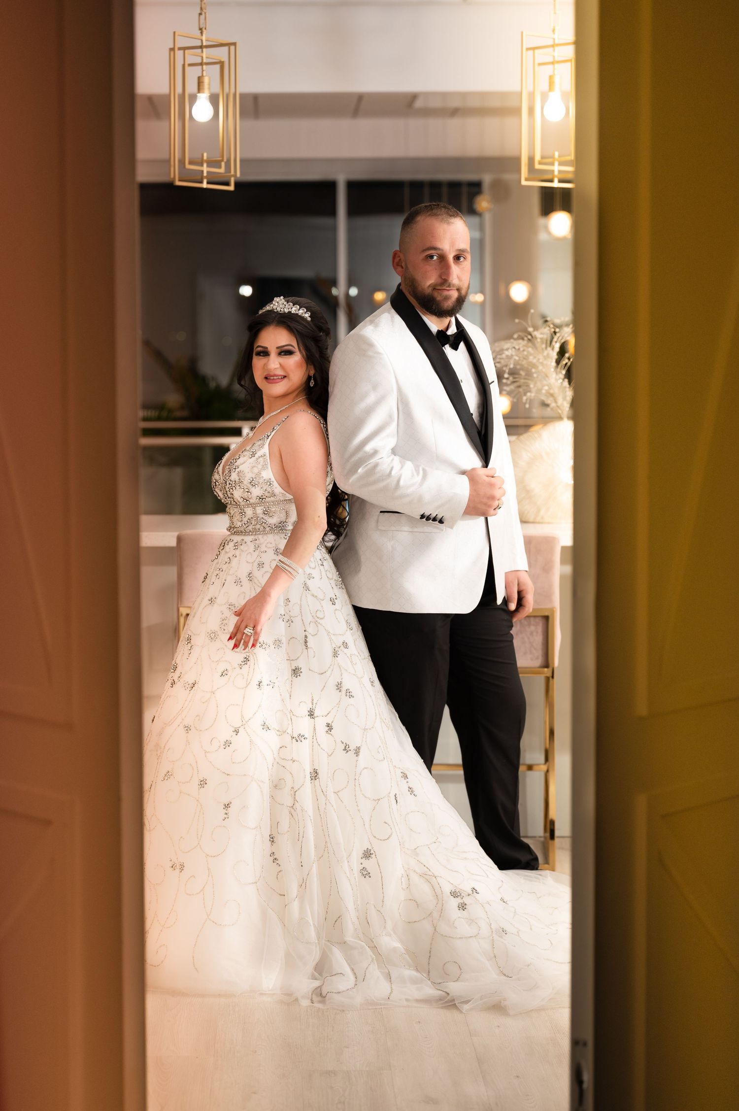 A glamorous couple poses in formal attire against modern gold lighting fixtures.